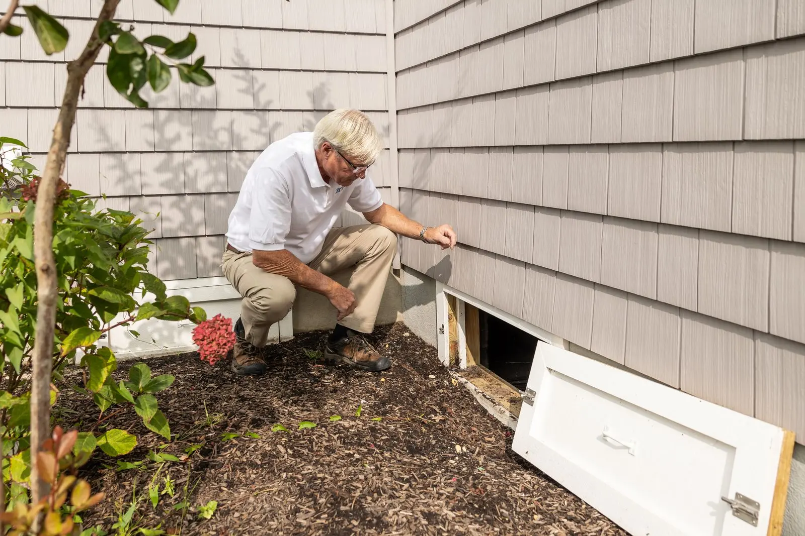 Home inspector kneeling outdoors to check a crawl space access beside a house